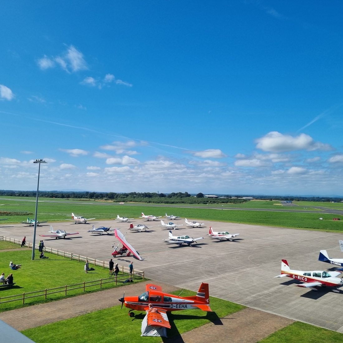 Aircrafts parked on a concrete apron at a regional airport, with people seated at picnic tables nearby,